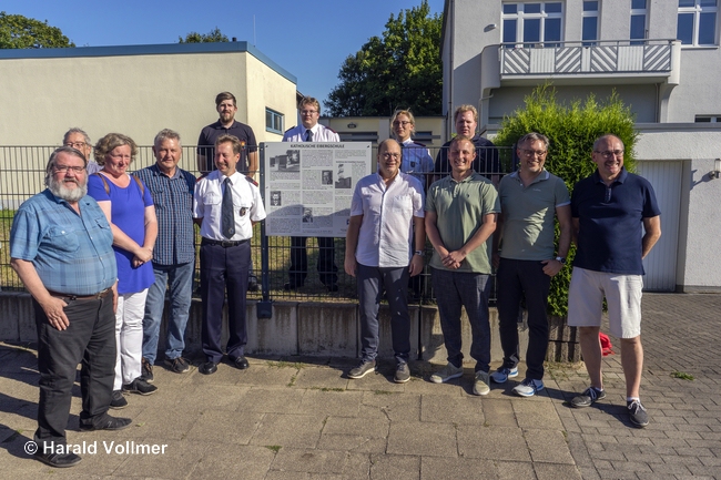 Freuen sich über die neue Denkmaltafel, v.links: Bezirksvertreter Ernst Potthoff, Klaus Geiser, Bezirksvertreterin Nicole Markner, 1. stellv. Bezirksbürgermeister Hagen Mertinat, Hauptbrandmeister René Schubert, Bezirksbürgermeister Frank Stienecker, Ratsherr Luca Ducrée, Bezirksvertreter David Herberg und Vorsitzender Christian Schlich; im Hintergrund Mitglieder der Löschgruppe Horst-Eiberg vor dem alten Schulgebäude rechts und dem heutigen Gerätehaus links.